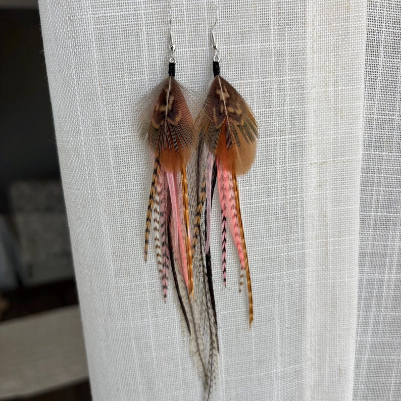 May include: A pair of feather earrings with a mix of brown, pink, and black feathers. The earrings have silver hooks and black accents. The feathers are arranged in a layered design, creating a flowing, bohemian look.