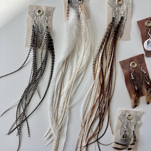 May include: A collection of feather earrings in various colors, including black and white speckled, solid white, and brown and tan. The earrings are displayed on small fabric cards with metal eyelets and a printed design.