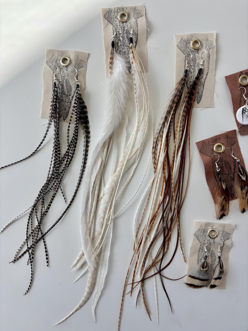 May include: A collection of feather earrings in various colors, including black and white speckled, solid white, and brown and tan. The earrings are displayed on small fabric cards with metal eyelets and a printed design.