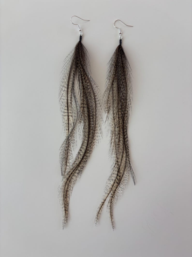 May include: A pair of long feather earrings. The earrings feature long, flowing feathers in shades of brown, gray, and white. The feathers are attached to silver-colored ear wires. The earrings are designed to dangle and sway.