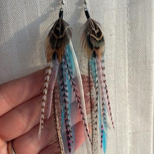 May include: A pair of feather earrings with silver hooks. The earrings feature a mix of brown, blue, pink, and white feathers. The feathers are arranged in a layered design, creating a flowing, eye-catching look. These earrings are a unique accessory.