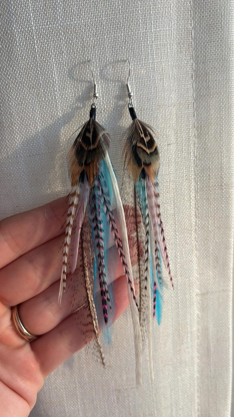 May include: A pair of feather earrings with silver hooks. The earrings feature a mix of brown, blue, pink, and white feathers. The feathers are arranged in a layered design, creating a flowing, eye-catching look. These earrings are a unique accessory.