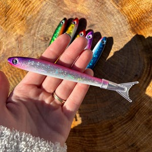 May include: A collection of colorful fishing lures in various shades of pink, purple, blue, green, and yellow. The lures are shaped like small fish with clear plastic tails. The lures are held against a wooden background.