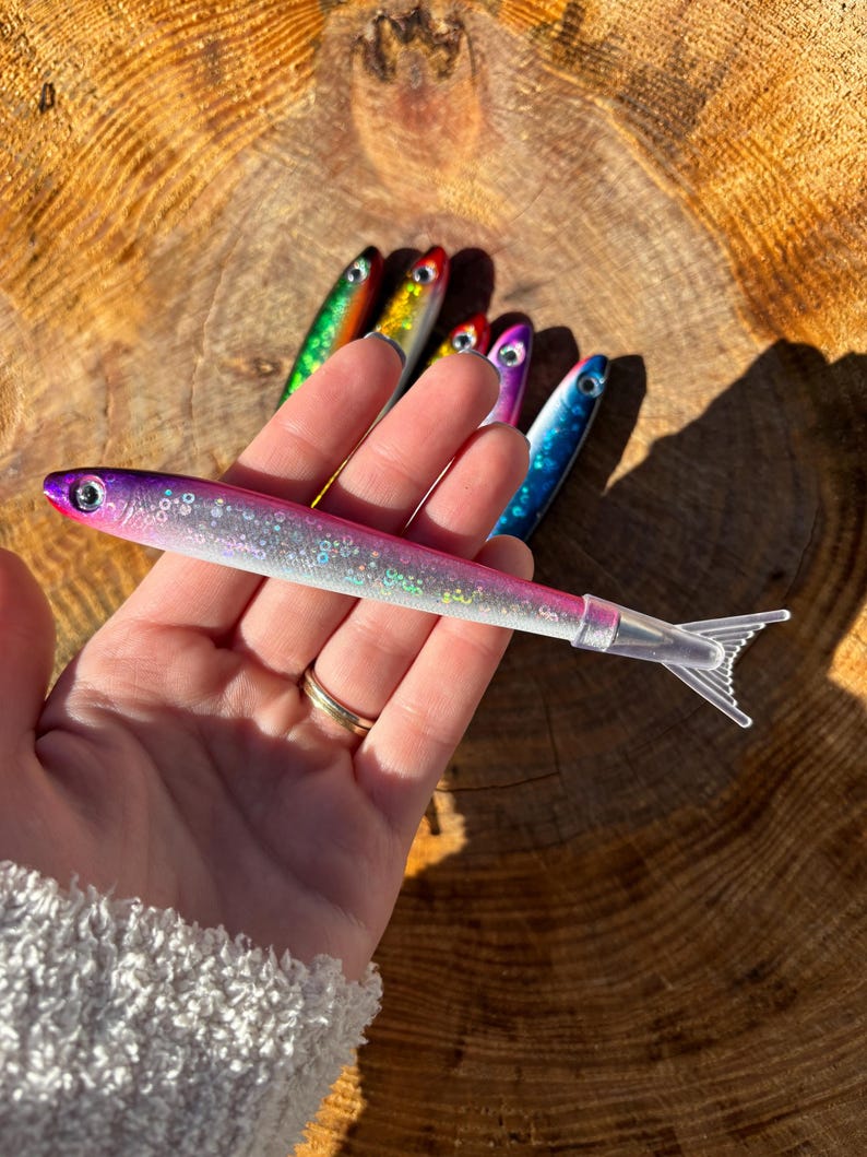 May include: A collection of colorful fishing lures in various shades of pink, purple, blue, green, and yellow. The lures are shaped like small fish with clear plastic tails. The lures are held against a wooden background.