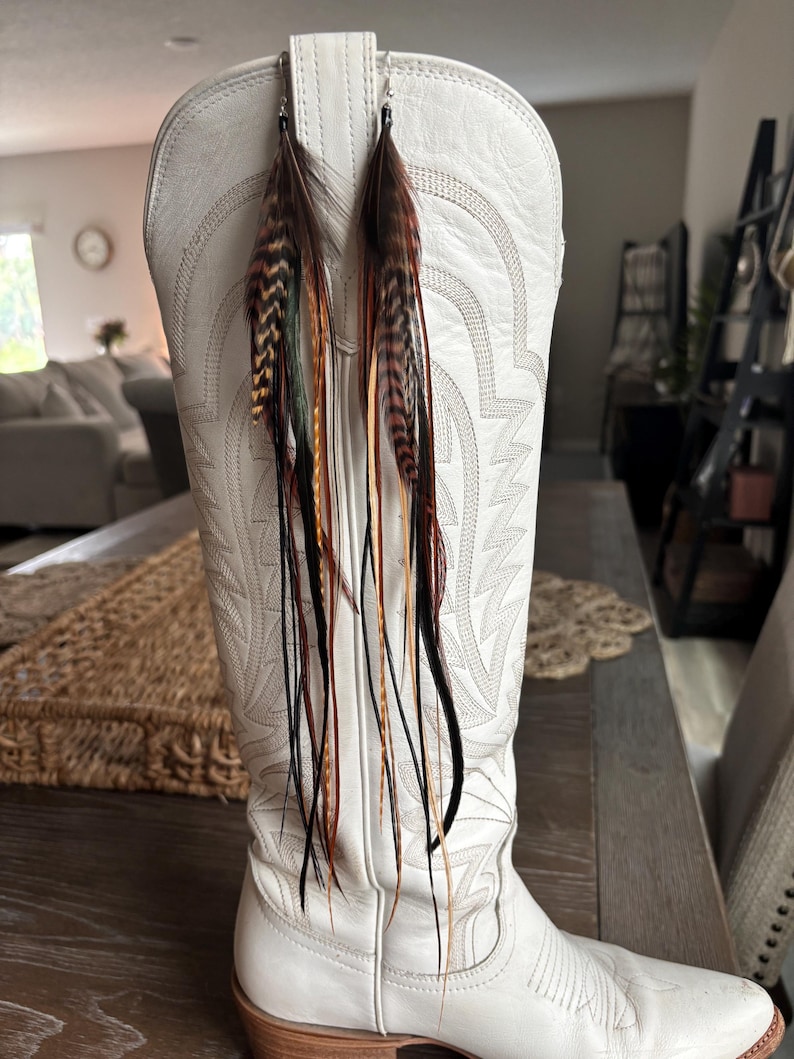May include: A pair of feather earrings with brown, black, and orange feathers hanging from silver hooks. The earrings are displayed on a white cowboy boot with decorative stitching.