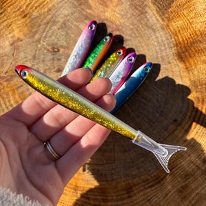 May include: A collection of five fish-shaped pens featuring glittery, multi-colored bodies with metallic finishes, realistic eyes, and clear plastic tail-shaped caps. Each pen displays a unique color gradient, including gold, purple, green, and blue.