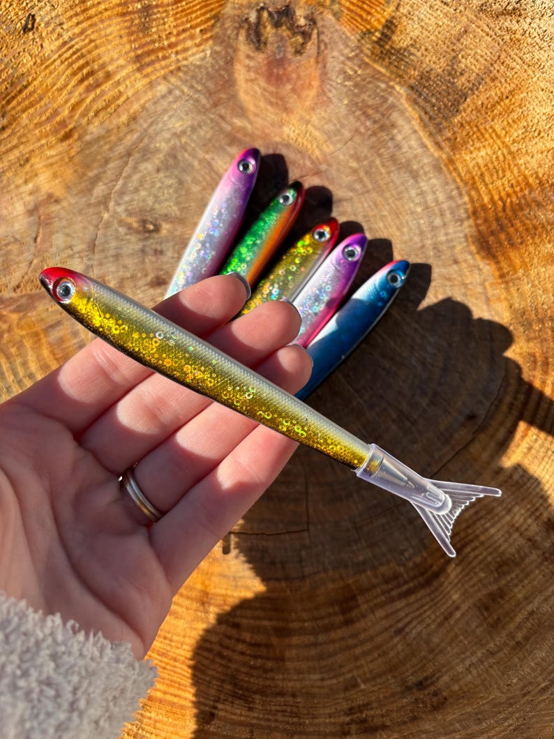 May include: A collection of five fish-shaped pens featuring glittery, multi-colored bodies with metallic finishes, realistic eyes, and clear plastic tail-shaped caps. Each pen displays a unique color gradient, including gold, purple, green, and blue.