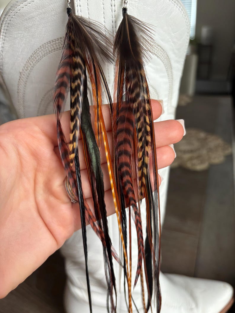 May include: Long feather earrings with a mix of brown, black, orange, and green feathers. The earrings are suspended from small silver-colored hooks. The feathers are long and flowing, creating a bohemian style.