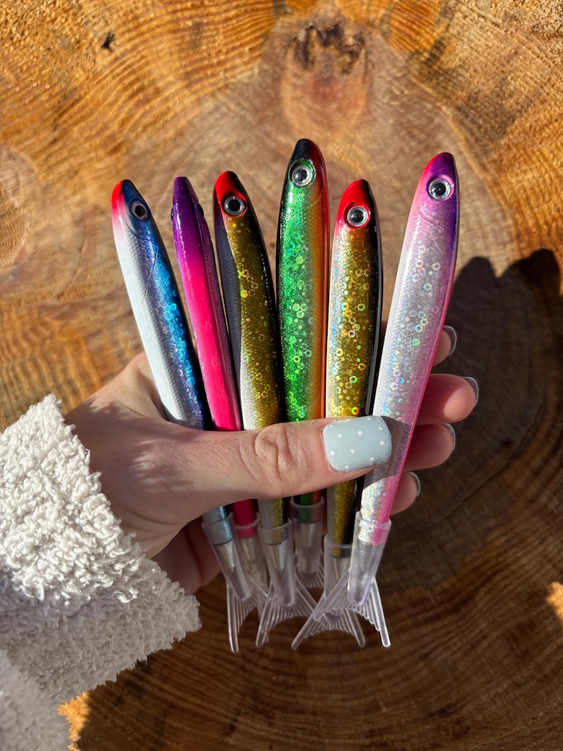 May include: A collection of colorful fishing lures in various shades of blue, pink, green, and gold. Each lure has a fish-like shape with a red or black eye and a clear plastic tail. The lures are held together.
