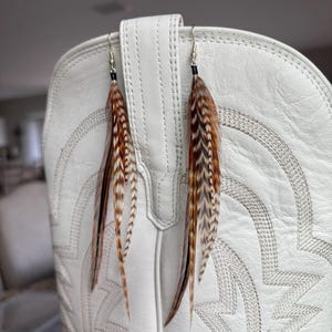 May include: A pair of feather earrings with brown, black, and white feathers hanging in front of a white cowboy boot. The earrings have silver-colored hooks and small black beads. The feathers are long and slender, with a natural, textured appearance.
