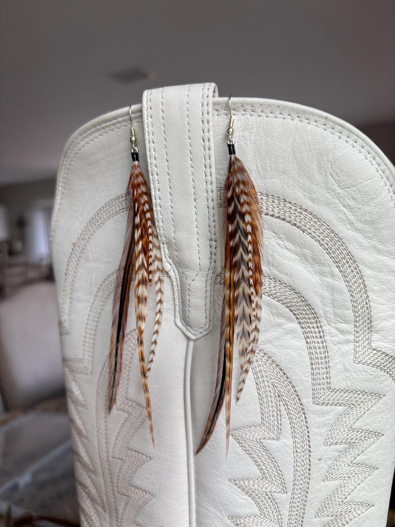 May include: A pair of feather earrings with brown, black, and white feathers hanging in front of a white cowboy boot. The earrings have silver-colored hooks and small black beads. The feathers are long and slender, with a natural, textured appearance.