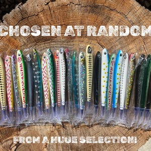 May include: A collection of fishing lures in various colors and patterns, displayed in clear plastic packaging. The lures are arranged on a wooden surface, with the text "CHOSEN AT RANDOM FROM A HUGE SELECTION!" in white.