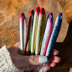 May include: A collection of colorful fishing lures. Each lure is shaped like a fish and features a vibrant color scheme, including silver, red, green, gold, pink, and blue. The lures have clear plastic fins and are held against a wooden background.