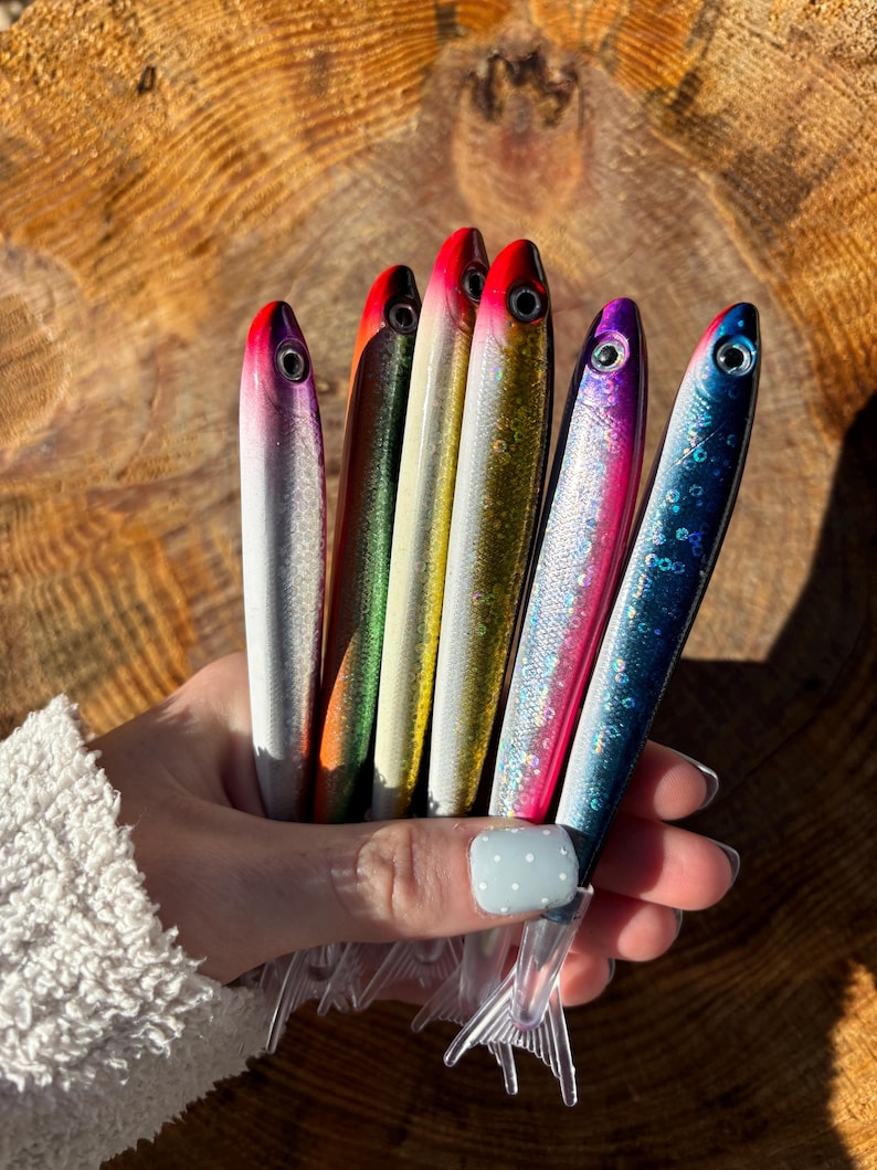 May include: A collection of colorful fishing lures. Each lure is shaped like a fish and features a vibrant color scheme, including silver, red, green, gold, pink, and blue. The lures have clear plastic fins and are held against a wooden background.