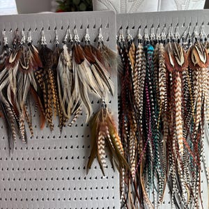 May include: A collection of feather earrings displayed on gray foam boards. The earrings feature a variety of colors, including brown, tan, black, and teal. The feathers are attached to silver-colored hooks.
