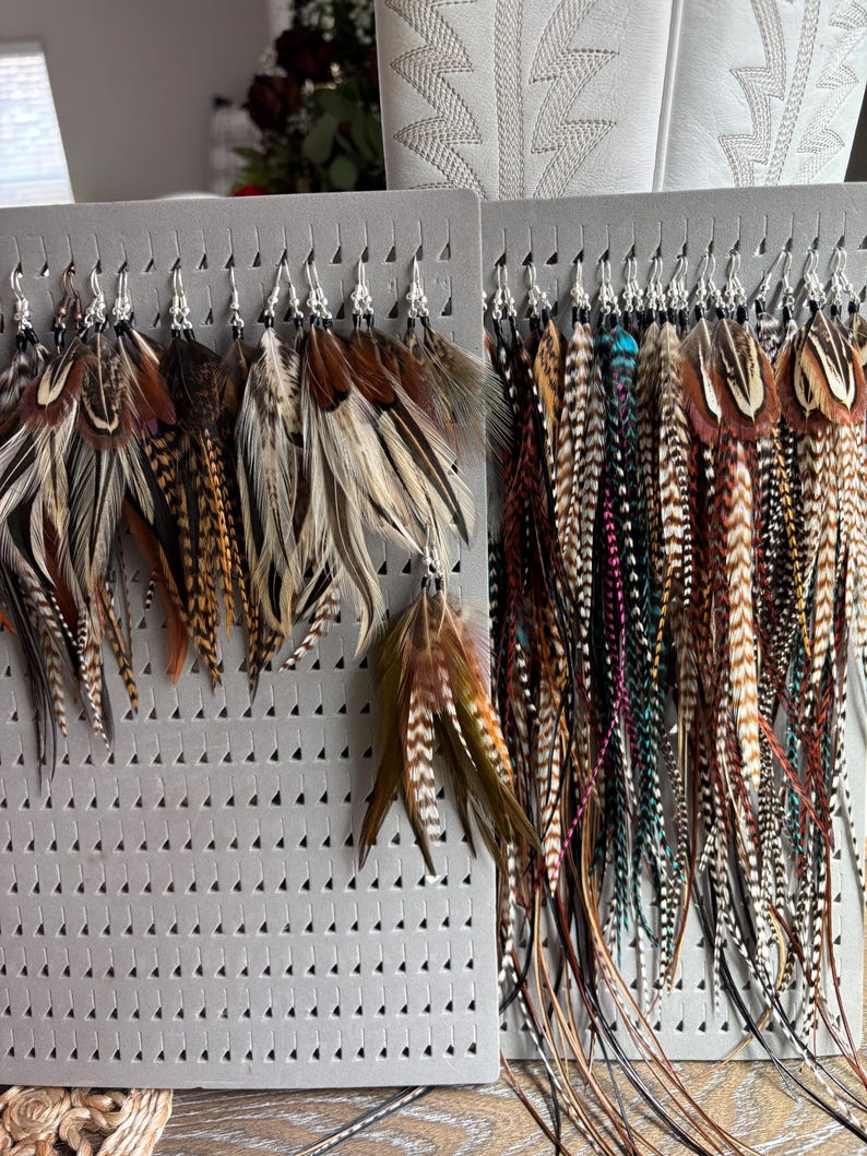 May include: A collection of feather earrings displayed on gray foam boards. The earrings feature a variety of colors, including brown, tan, black, and teal. The feathers are attached to silver-colored hooks.