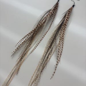 May include: A pair of long feather earrings. The earrings feature a mix of brown, gray, and cream-colored feathers. Each earring has a silver-colored hook. The feathers are arranged to create a flowing, elongated design.