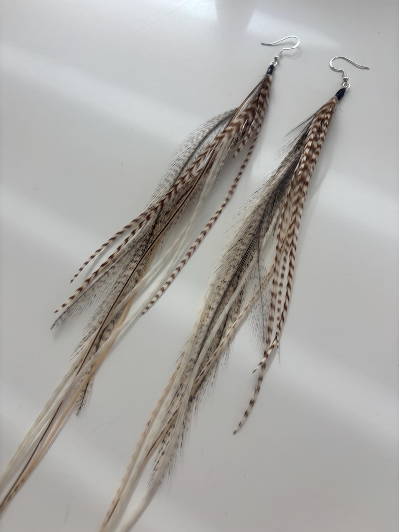 May include: A pair of long feather earrings. The earrings feature a mix of brown, gray, and cream-colored feathers. Each earring has a silver-colored hook. The feathers are arranged to create a flowing, elongated design.