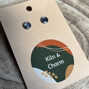 Może przedstawiać: Para opalizujących kolczyków sztyftów na beżowej karcie z okrągłą naklejką. Naklejka zawiera biały tekst "Kiln & Charm" na ciemnozielonym tle, z brązowym i białym wzorem. Kolczyki są małe i okrągłe.