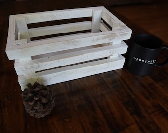 Peut inclure: Une caisse en bois blanche avec une finition vieillie. La caisse est vide et repose sur une surface en bois foncé. Il y a un cône de pin et une tasse à café noire avec le texte "The Porches" dessus.