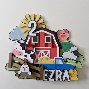 May include: A farm-themed papercraft decoration with a red barn, blue truck, and farm animals. The design includes a cow, pig, sheep, and chicken, with the name "Ezra" and the number "2". A yellow sun and hay bale complete the scene, suitable for a child's birthday.