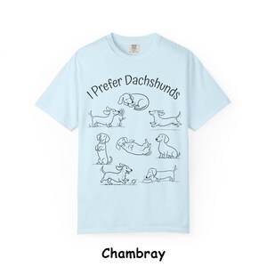 Pode incluir: Uma t-shirt azul-clara com um gr&aacute;fico de linhas pretas de sete c&atilde;es dachshund em v&aacute;rias posi&ccedil;&otilde;es e o texto "I Prefer Dachshunds" arqueado sobre os c&atilde;es. A t-shirt tem mangas curtas, gola redonda e um design de corte padr&atilde;o.