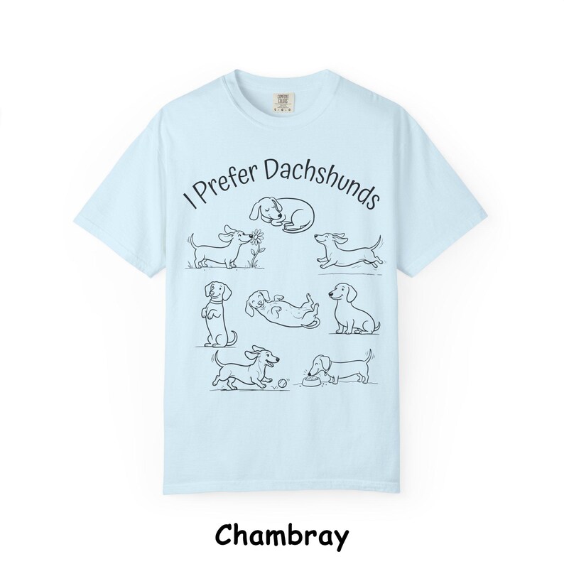 Pode incluir: Uma t-shirt azul-clara com um gr&aacute;fico de linhas pretas de sete c&atilde;es dachshund em v&aacute;rias posi&ccedil;&otilde;es e o texto "I Prefer Dachshunds" arqueado sobre os c&atilde;es. A t-shirt tem mangas curtas, gola redonda e um design de corte padr&atilde;o.