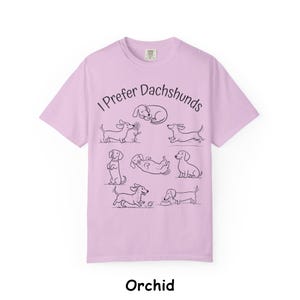 Pode incluir: Uma t-shirt roxo-claro com o texto preto I Prefer Dachshunds e uma s&eacute;rie de ilustra&ccedil;&otilde;es de linhas pretas representando v&aacute;rios c&atilde;es salsicha em diferentes poses, incluindo a dormir, a correr, sentado e a brincar com uma bola.