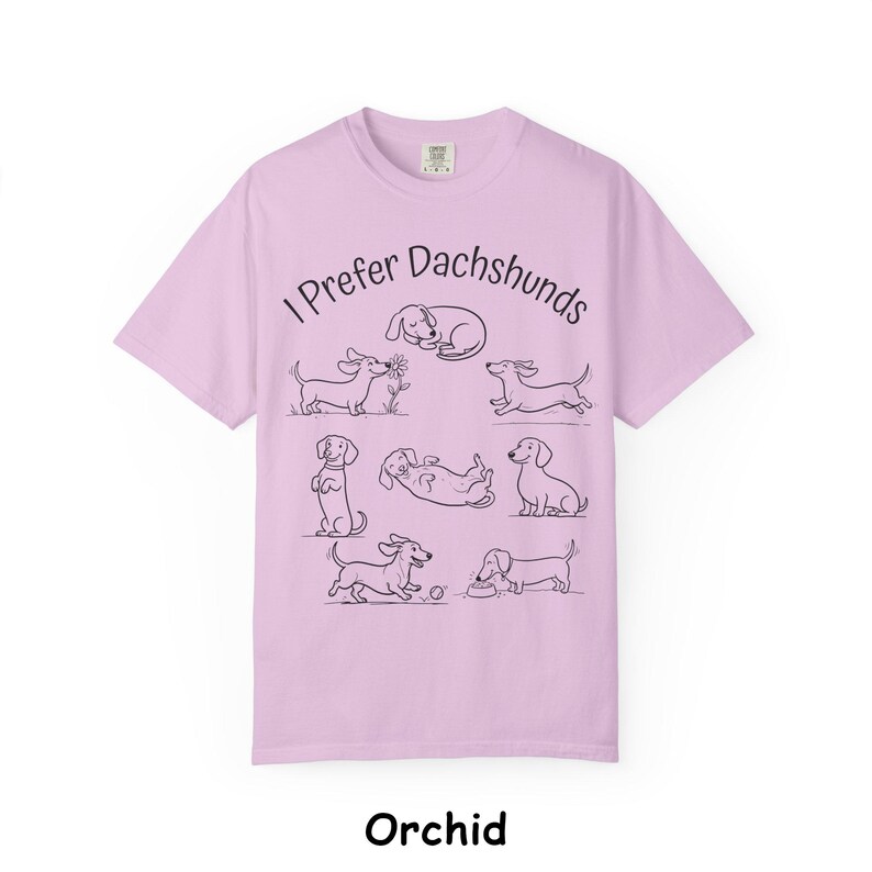 Pode incluir: Uma t-shirt roxo-claro com o texto preto I Prefer Dachshunds e uma s&eacute;rie de ilustra&ccedil;&otilde;es de linhas pretas representando v&aacute;rios c&atilde;es salsicha em diferentes poses, incluindo a dormir, a correr, sentado e a brincar com uma bola.