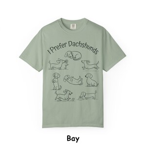Pode incluir: Uma t-shirt de manga curta verde s&aacute;lvia com um gr&aacute;fico de linhas pretas de v&aacute;rios c&atilde;es dachshund em diferentes posi&ccedil;&otilde;es. O texto "I Prefer Dachshunds" est&aacute; impresso numa fonte preta simples acima das ilustra&ccedil;&otilde;es dos c&atilde;es a brincar.