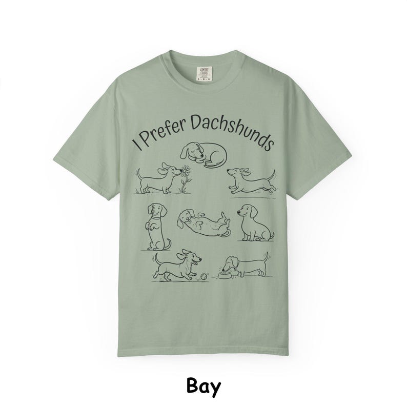 Pode incluir: Uma t-shirt de manga curta verde s&aacute;lvia com um gr&aacute;fico de linhas pretas de v&aacute;rios c&atilde;es dachshund em diferentes posi&ccedil;&otilde;es. O texto "I Prefer Dachshunds" est&aacute; impresso numa fonte preta simples acima das ilustra&ccedil;&otilde;es dos c&atilde;es a brincar.
