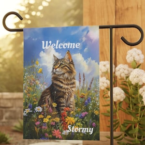 May include: A garden flag featuring a detailed illustration of a cat amidst a field of colorful wildflowers under a blue sky. The word "Welcome" is at the top, and "Stormy" is at the bottom.