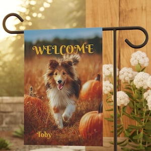 Può includere: Una bandiera da giardino con un cane che corre attraverso un campo di zucche. La parola "WELCOME" è scritta in oro, con il nome del cane "Toby" in basso. La bandiera è montata su un supporto di metallo nero.