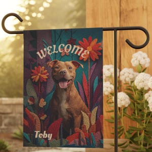 May include: A garden flag featuring a smiling brown dog with the name "Toby" below the image. The word "welcome" is at the top. The background includes colorful flowers and foliage. The flag is displayed on a black metal stand.