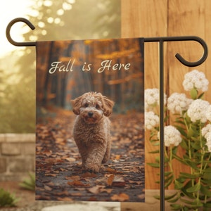 Puede incluir: Una bandera de jardín con un perro marrón y blanco caminando por un sendero de hojas de otoño. El texto "Fall is Here" está impreso en la bandera.