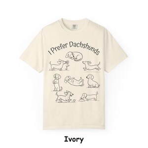 Pode incluir: Uma t-shirt de manga curta na cor marfim com um gr&aacute;fico de linhas pretas de v&aacute;rios c&atilde;es dachshund em diferentes poses. O texto "I Prefer Dachshunds" est&aacute; impresso numa fonte preta simples acima das ilustra&ccedil;&otilde;es dos c&atilde;es a brincar.
