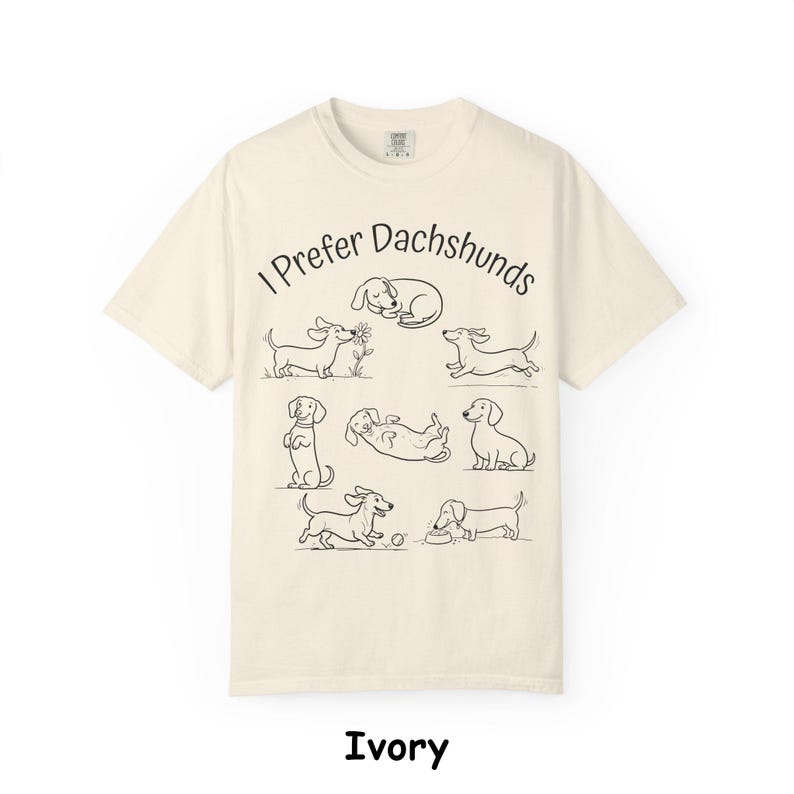 Pode incluir: Uma t-shirt de manga curta na cor marfim com um gr&aacute;fico de linhas pretas de v&aacute;rios c&atilde;es dachshund em diferentes poses. O texto "I Prefer Dachshunds" est&aacute; impresso numa fonte preta simples acima das ilustra&ccedil;&otilde;es dos c&atilde;es a brincar.