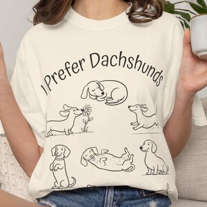 Pode incluir: Uma t-shirt de manga curta em tom off-white com o texto preto I Prefer Dachshunds e v&aacute;rias ilustra&ccedil;&otilde;es de linhas pretas de c&atilde;es dachshund em v&aacute;rias poses, incluindo a dormir, a cheirar uma flor, a correr, sentado e deitado de costas.