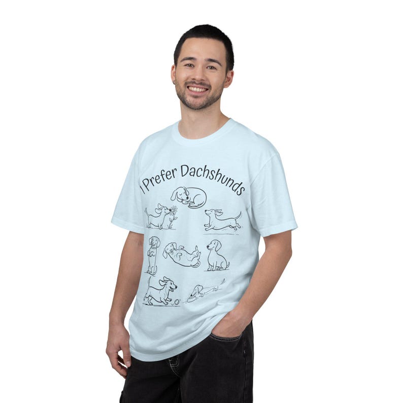 Pode incluir: Uma t-shirt de manga curta azul-clara com o texto preto "I Prefer Dachshunds" arqueado sobre uma cole&ccedil;&atilde;o de sete ilustra&ccedil;&otilde;es de linhas pretas de c&atilde;es dachshund em v&aacute;rias poses, incluindo a dormir, a correr e a brincar.