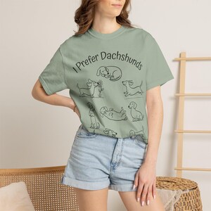 Pode incluir: Uma t-shirt de manga curta verde-s&aacute;lvia com ilustra&ccedil;&otilde;es de linhas pretas de v&aacute;rios c&atilde;es dachshund em diferentes poses e o texto "I Prefer Dachshunds" impresso numa fonte preta simples na parte superior do peito da pe&ccedil;a.