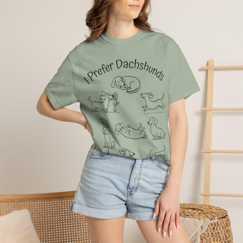 Pode incluir: Uma t-shirt de manga curta verde-s&aacute;lvia com ilustra&ccedil;&otilde;es de linhas pretas de v&aacute;rios c&atilde;es dachshund em diferentes poses e o texto "I Prefer Dachshunds" impresso numa fonte preta simples na parte superior do peito da pe&ccedil;a.