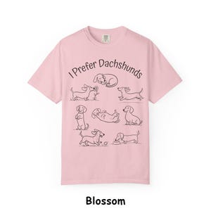 Pode incluir: Uma t-shirt de manga curta em rosa claro com o texto preto I Prefer Dachshunds e uma s&eacute;rie de ilustra&ccedil;&otilde;es de linhas pretas que retratam v&aacute;rios c&atilde;es dachshund em diferentes poses, incluindo a dormir, a correr, a sentar e a brincar.
