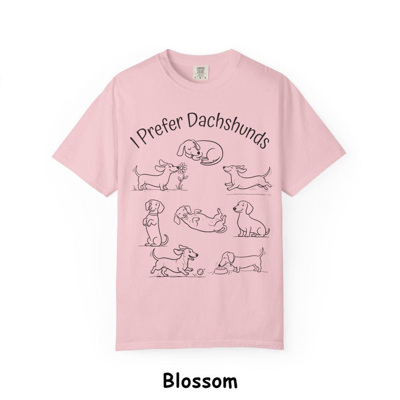 Pode incluir: Uma t-shirt de manga curta em rosa claro com o texto preto I Prefer Dachshunds e uma s&eacute;rie de ilustra&ccedil;&otilde;es de linhas pretas que retratam v&aacute;rios c&atilde;es dachshund em diferentes poses, incluindo a dormir, a correr, a sentar e a brincar.