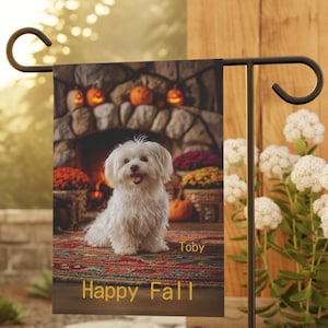 May include: A garden flag featuring a white dog named Toby sitting in front of a fireplace decorated for fall. The flag has the words "Happy Fall" in gold lettering.