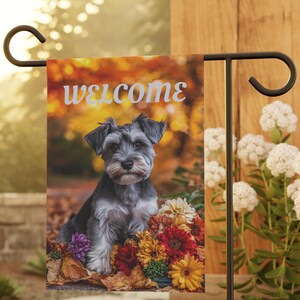 Puede incluir: Una bandera de jardín de bienvenida con un schnauzer negro y gris sentado en un lecho de flores de colores otoñales. La bandera tiene un fondo borroso de follaje otoñal y la palabra "bienvenido" en texto blanco.