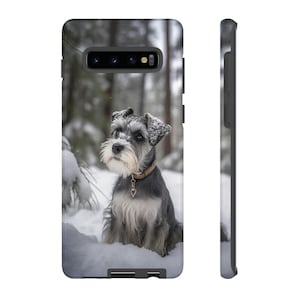Könnte beinhalten: Ein kleiner, grau-weißer Zwergschnauzer sitzt im Schnee. Der Hund trägt ein braunes Halsband mit einem goldenen Herzanhänger. Der Hund schaut in die Kamera.