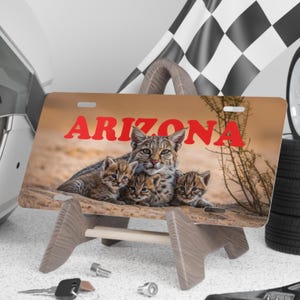 K&ouml;nnte beinhalten: Kennzeichen mit dem Wort "ARIZONA" in Rot, das eine Familie von Rotluchsen zeigt. Das Schild wird auf einem Holzst&auml;nder pr&auml;sentiert, mit einer Rennflagge und Reifen im Hintergrund. Die Rotluchse sind braun und beige mit schwarzen Flecken.