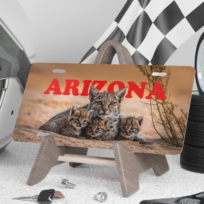 K&ouml;nnte beinhalten: Kennzeichen mit dem Wort "ARIZONA" in Rot, das eine Familie von Rotluchsen zeigt. Das Schild wird auf einem Holzst&auml;nder pr&auml;sentiert, mit einer Rennflagge und Reifen im Hintergrund. Die Rotluchse sind braun und beige mit schwarzen Flecken.