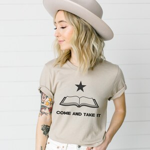 May include: Tan-colored t-shirt featuring a black star above an open book graphic. The text "COME AND TAKE IT" is printed below the book. The shirt is a short-sleeved crew neck.