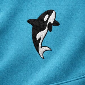 May include: A turquoise sweatshirt featuring an embroidered orca whale design. The orca is black and white, with the whale's body in black and the belly and accents in white. The sweatshirt is made of a soft, comfortable material.